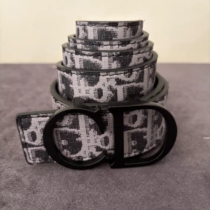 Svartvit Dior bälte med monogram - Snyggt bälte från Dior i svart och vitt med tydligt monogrammönster och stor svart CD-spänne. Bältet har en modern vibe och är gjort i ett tåligt material med struktur. Perfekt för att lyfta vilken outfit som helst med en lyxig touch.