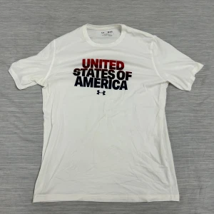 Vit Under Armour t-shirt USA-print - Vit t-shirt från Under Armour med stort tryck 'United States of America' i rött och svart på bröstet. Klassisk rund hals, korta ärmar och loose fit. Perfekt för dig som gillar sportig och avslappnad stil.