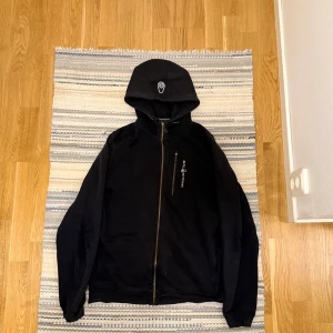 Svart hoodiejacka från Sail Racing - Svart hoodiejacka från Sail Racing med dragkedja framtill och bröstficka med logga. Jackan har huva med broderad detalj och är tillverkad i mjuk bomullsmix. Perfekt för chill dagar och streetwear-vibe. Den har varsin skavank på varsin armbåge