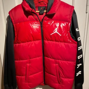 Västjacka från Jordan med logga - Säljer en röd dunväst från Jordan med klassisk Jumpman-logga i vitt på bröstet. Västen har dragkedja framtill, två fickor och hög krage. Perfekt för lager-på-lager och streetwear-stil. Materialet är glansigt och känns sportigt.