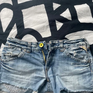 Replay jeansshorts med slitningar - Snygga ljusblå jeansshorts från Replay med slitna detaljer, råa kanter och gula dragkedjor på bakfickorna. Klassisk femficksmodell i 100% bomull med coolt Replay-läderpatch bak. Perfekta för en avslappnad streetstil. Säljer för att dom inte passar, som ni ser på sista bilden står det storlek, jag passade i dom och bär S