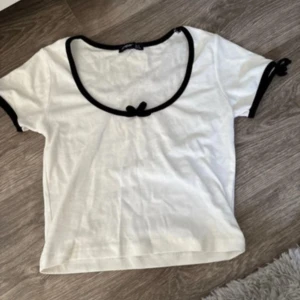 Vit topp med svarta rosetter - Supersöt vit topp med svarta kanter och små rosetter vid halsringningen och ärmsluten. Rundad, djup urringning och korta ärmar. Perfekt för en trendig och söt look.