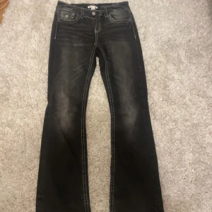 Svarta/gråabootcut jeans från Gina Tricot - Svarta/gråa jeans från Gina Tricot med bootcut-modell och markerade kontrastsömmar. Klassisk femficksdesign med snygga detaljer på bakfickorna och dragkedja framtill. Perfekta för dig som gillar en retroinspirerad look.