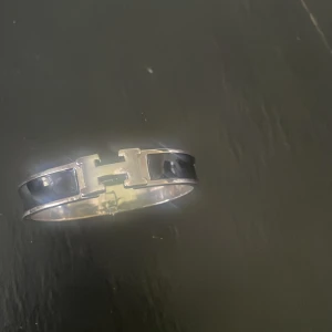 Svart armband med H-detalj - Snyggt armband i silverfärgad metall med svart emalj och den ikoniska H-detaljen framtill. Stilren och lyxig accessoar som lyfter vilken outfit som helst.