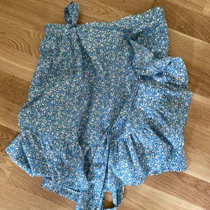 Blommig blå omlottkjol med volang - Supersöt kort blå kjol med vitt blommönster och volangkant. Omlottmodell med knyt i midjan, perfekt för en fräsch och somrig look. Lätt och luftig, passar perfekt till sneakers eller sandaler.