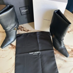 Saint Laurent Stövletter - Exklusiva svarta stövletter från Saint Laurent i slätt skinn med spetsig tå och hög klack. Snyggt YSL-emblem i guld vid ankeln och elegant, minimalistisk design. Perfekta för dig som vill sticka ut med lyxiga detaljer och tidlös stil.