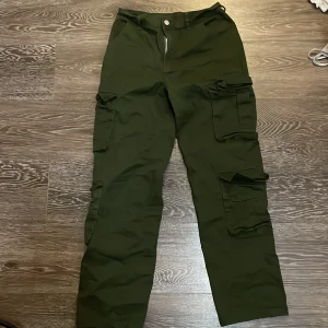 Mörkgröna cargopants med fickor - Säljer ett par mörkgröna cargopants med flera stora fickor på benen och klassisk rak passform. Byxorna har dragkedja och knapp framtill och är tillverkade i ett slitstarkt bomullstyg. Perfekta för en avslappnad streetstil. Bara använd 1-3 gånger 