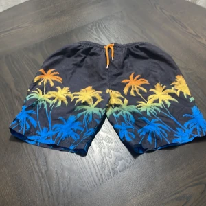 Svarta badshorts med palmmönster Primark - Coola svarta badshorts från Primark med färgglada palmer i blått, grönt, gult och orange längs benen. Shortsen har elastisk midja med orange snörning och meshfoder på insidan. Perfekta för stranden eller poolen!