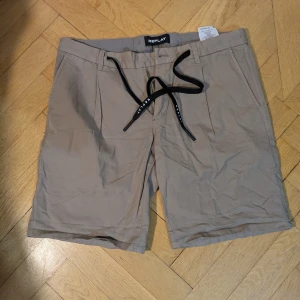 Beige shorts från Replay med snörning - Snygga beige shorts från Replay med svarta snören i midjan och flera fickor, både fram och bak. Klassisk passform och stilren design, perfekta för varma dagar. Tillverkade i mjuk bomull och linne som känns skön mot huden. Diskreta Replay-detaljer och knappstängning bak. Knapp saknas fram, men du kan använda snörena eller sy dit en egen knapp. Lite skrynkliga men i bra skick. Tillståndet sätts i bra skick pga knappen, men annars i mycket bra skick. 