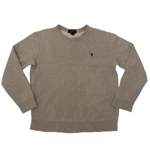 Grå sweatshirt från Polo Ralph Lauren - En stilren grå sweatshirt från Polo Ralph Lauren med klassisk rund hals och broderad logga på bröstet. Tröjan har långa ärmar och är tillverkad i mjuk bomullsmix som är perfekt för chill dagar. Enkel och clean look som funkar till allt.