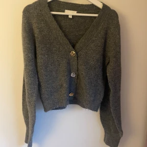 Grå kofta från H&M med guldknappar - Mysig grå kofta från H&M i stickat material. Koftan har v-ringning, långa ärmar och tre snygga guldiga knappar framtill som ger en cool touch. Perfekt att slänga på sig när det är kyligt ute.