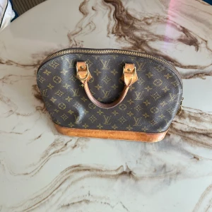Louis Vuitton Monogram handväska - Säljer en klassisk Louis Vuitton handväska i brunt monogrammönster med ikoniska LV-loggor. Väskan har rundad form, detaljer i ljust skinn och mässingsfärgade metalldelar. Dragkedja upptill och lås med Louis Vuitton-gravyr. Perfekt för dig som gillar lyxig och tidlös stil.