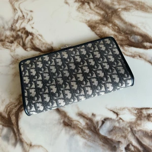 Christian Dior monogram plånbok - Snygg plånbok från Christian Dior med klassiskt monogrammönster i svart och beige. Rektangulär form med metallspänne där Dior-loggan syns. Utsidan är i textil med tryckt mönster och insidan i svart skinn. Perfekt för dig som gillar lyxiga accessoarer.