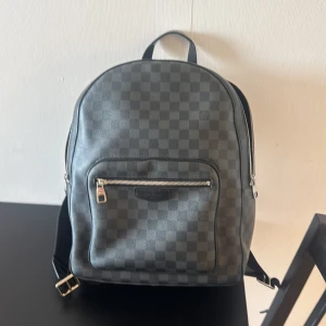 Louis Vuitton Damier Graphite ryggsäck - Säljer en snygg Louis Vuitton ryggsäck i klassiska Damier Graphite-mönstret. Väskan är svart och grå med rutigt mönster, har justerbara axelremmar, dragkedja framtill och detaljer i skinn. Perfekt för dig som vill ha en lyxig och stilren väska till vardagen. Kvitto från butiken jag köpt den på finns att få via mail.