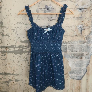 Mörkblå for love & lemons blommig kortklänning med volang - Supersöt mörkblå kortklänning med små blommor i lila och vitt. Klänningen har smockad midja, volangiga axelband och en liten rosett framtill. Perfekt för sommarens alla äventyr och har en romantisk vibe.