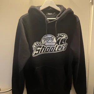 Svart Trapstar hoodie London Shooters - Svart hoodie från Trapstar med fett London Shooters-tryck framtill. Klassisk huva med snörning och känguruficka. Materialet känns mjukt och håller värmen bra.