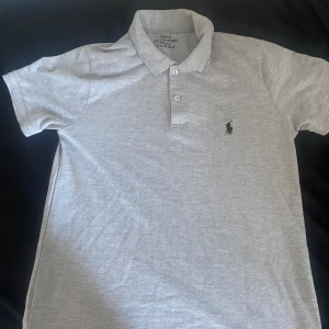 Grå pikétröja från Ralph Lauren - Klassisk grå pikétröja från Ralph Lauren med korta ärmar och broderad logga på bröstet. Tröjan har krage och två knappar framtill. Perfekt basic till garderoben och funkar året runt. Jag glömde skriva innan men priset är fast.