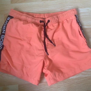Orange Björn Borg badshorts XS - Snygga orange badshorts från Björn Borg i storlek XS. De har svart sidopanel med vit logga, elastisk midja och snörning framtill. Perfekta för stranden eller poolen, med meshfoder för extra komfort.