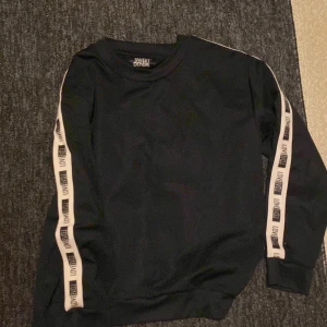 Svart sweatshirt från Sweet Denim - Svart sweatshirt från Sweet Denim med vita band längs ärmarna där det står 'DENIM' i svart text. Tröjan har rund halsringning och långa ärmar, perfekt för en avslappnad streetstyle-look.