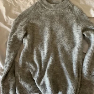 Grå stickad tröja med hög krage - Mysig grå stickad tröja med högre krage och långa ärmar. Tröjan har en ribbad struktur och är perfekt för kyliga dagar. Enkel och stilren design som passar till det mesta.