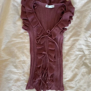 Plommonlila volangtopp från Zara - Supersöt plommonlila topp från Zara med volanger längs v-ringningen och knyt framtill. Ribbad struktur och ärmlös design som ger en trendig och feminin look. Perfekt att styla med jeans eller kjol för en cool vibe. Nyskick!