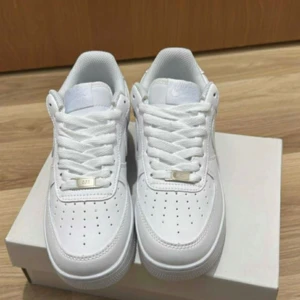 Nike Air Force 1 helvita sneakers - Klassiska Nike Air Force 1 sneakers i helvitt skinn med perforerad tå, vit sula och ikonisk Swoosh på sidan. Lågt skaft, snörning och diskreta detaljer i silver. Perfekta för dig som vill ha en clean och tidlös look. Aldrig använda bara testade fick i present 