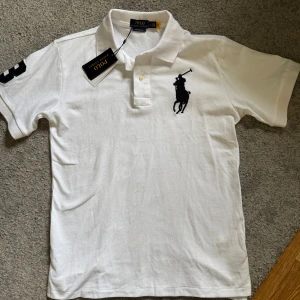 Vit pikétröja från Polo Ralph Lauren - Klassisk vit pikétröja från Polo Ralph Lauren med svart broderad logga på bröstet och nummerdetalj på ärmen. Tröjan har krage, knappar framtill och är tillverkad i mjuk bomull. Perfekt för en clean och sportig stil.