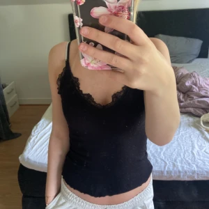 Svart linnetopp med spetskant - Säljer en svart linnetopp med smala axelband och snygg spetskant längs v-ringningen. Toppen är croppad och har en feminin, enkel stil som passar perfekt till jeans eller kjol. Superenkel att matcha och riktigt skön att bära.