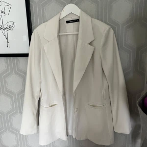 Vit kavaj från Gina Tricot - Stilren vit kavaj från Gina Tricot i klassisk modell med spetsiga slag och två fickor framtill. Kavajen har knäppning med två knappar och är helfodrad. Perfekt för en clean och fräsch look. Oversize look.