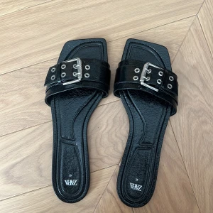 Svarta sandaler med spänne från Zara - Svarta sandaler från Zara med och silverfärgat spänne med ett snyggt glansigt material. Säljer pågrund av att de är för stora för mig… och använt skorna endast en gång.