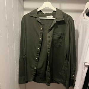 Mörkgrön skjorta från Fast West - Stilren mörkgrön skjorta från Fast West med klassisk krage, bröstficka och knäppning framtill. Skjortan har lång ärm och en avslappnad passform, perfekt att bära öppen eller stängd. Tillverkad i mjuk bomull för en skön känsla.