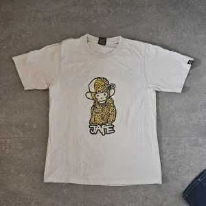 Vit JAPE t-shirt med leopardapa - Vit t-shirt från JAPE med coolt tryck av en apa i leopardmönstrad hoodie och keps på framsidan. Baksidan har en grafisk JAPE-logga och liten apfigur nere till vänster. Klassisk passform och rund hals. Perfekt för dig som gillar streetwear med unik twist.