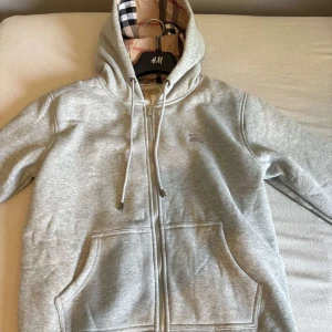 Grå hoodie från Burberry med dragkedja - Snygg grå hoodie från Burberry med dragkedja, huva med snörning och klassisk logga broderad på bröstet. Mjuk bomullsblandning och två stora fickor(pris går att diskutera)