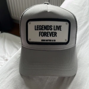 Grå keps John Hatter & Co - Snygg grå keps från John Hatter & Co med svart mesh baktill och böjd skärm. Framtill sitter en vit skylt med texten 'Legends Live Forever' i svart. Kepsen har justerbar passform och cool streetstyle-vibe.