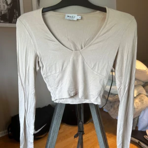 Beige croppad topp från NA-KD - Säljer en beige croppad topp från NA-KD med lång ärm och v-ringning. Toppens sömmar ger en snygg formad look och den är tillverkad i ett mjukt, stretchigt material som känns skönt mot huden. Perfekt att styla med höga jeans eller kjol. 🩷