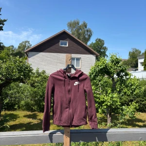 Vinröd hoodiejacka från Nike - Säljer en vinröd hoodiejacka från Nike med dragkedja framtill och huva. Jackan har en klassisk sportig look med Nike-logga på bröstet och är tillverkad i mjuk bomullsmix. 