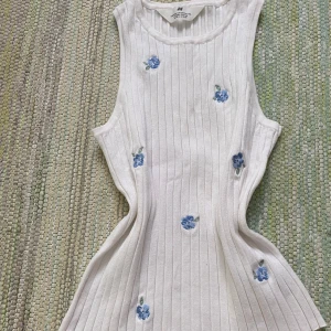 Vit ribbad klänning med blå blommor H&M - Superfin vit ribbad kortklänning från H&M med broderade blå blommor. Ärmlös modell med rund halsringning och figurnära passform. Perfekt för en fräsch och somrig look!