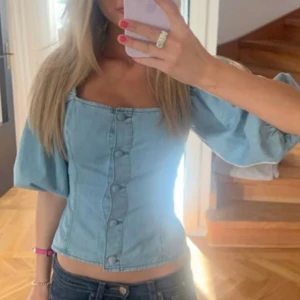 Ljusblå jeanskorsett med puffärmar - Trendig ljusblå korsettopp i jeanstyg med markerad midja och stora puffärmar. Toppen har fyrkantig urringning och knappar framtill som ger en snygg detalj. Perfekt för dig som vill sticka ut med en cool och feminin look.