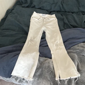 Vita bootcut jeans från Zara, strl 34 - Snygga vita bootcut jeans från Zara i storlek 34. Byxorna har slitna detaljer vid knäna och fransade benslut för en trendig look. Används några gånger men den är ändå i bra skick. Den är lågmidjad och perfekt för dig som letar efter vita jeans med ett billigt pris, Perfekt för sommaren! 
