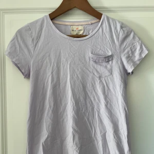 Ljuslila t-shirt Holly & Whyte XS - Supersöt ljuslila t-shirt från Holly & Whyte med rund halsringning och liten bröstficka med volangdetalj. Skön och mjuk i bomull, perfekt till jeans eller kjol. Enkel och stilren design som passar till många olika looks.