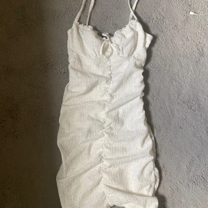 Vit kort sommarklänning - Supersöt vit kort klänning med smala axelband, broderade detaljer och knyt framtill. Klänningen har en feminin passform med rynkade sömmar längs mitten och är perfekt för varma dagar.