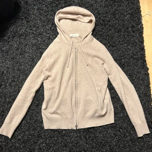 Beige hoodie i kashmir från Sayless - Mjuk och stilren beige hoodie från Sayless i kashmir. Tröjan har dragkedja framtill, huva med snörning och två fickor. Perfekt för dig som gillar en clean look och vill ha något riktigt skönt och lyxigt till vardags. 