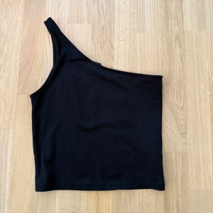 Svart oneshoulder topp Gina Tricot - Snygg svart ribbad topp från Gina Tricot med oneshoulder-design. Toppen är croppad och har en tight passform som framhäver axlarna. Perfekt för dig som vill ha en stilren och trendig look. Materialet är mjukt och stretchigt för extra komfort. Storlek s