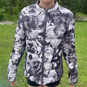 Blommig svartvit pufferjacka - Säljer en riktigt snygg pufferjacka med svartvitt blommönster. Jackan har dragkedja framtill, hög krage och långa ärmar. Perfekt för dig som vill sticka ut med en unik stil. Jackan är vadderad och passar bra när det är kyligt ute.                                                  Jackan har även fickor 