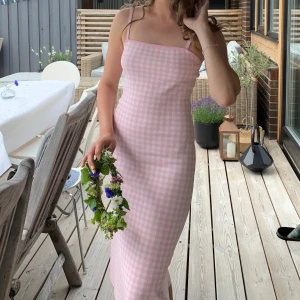 Rosa rutig långklänning med smala axelband - Supersöt långklänning i ljusrosa och vitt rutigt mönster. Klänningen har smala axelband och rak halsringning, sitter figurnära och har en klassisk somrig vibe. Perfekt för dig som gillar en feminin och trendig stil.
