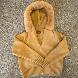 Beige cardigan jacka med fuskpäls, Alessa faux fur  - Mysig beige cardigan jacka från Abercrombie med huva kantad av fuskpäls. Jackan är stickad, har dragkedja framtill och ribbade muddar vid ärmslut och nederkant. Perfekt för kyliga dagar och ger en chill vibe.