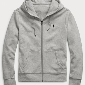 Grå hoodie från Polo Ralph Lauren - Grå hoodie från Polo Ralph Lauren med dragkedja, huva med snörning och klassisk broderad logga på bröstet. Tillverkad i mjuk bomullsmix, har två fickor framtill och ribbade muddar. Perfekt för en chill och stilren look. Priset kan självklart diskuteras!