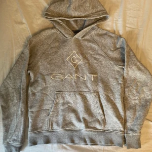 Grå hoodie från GANT med broderad logo - Säljer en grå hoodie från GANT med broderad vit logo på bröstet. Tröjan har huva, känguruficka och långa ärmar. Perfekt för dig som gillar en clean och klassisk stil. Mjuk och skön bomullsblandning. 