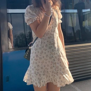 Blommig kortklänning från NA-KD - Supersöt kortklänning från NA-KD i ljusbeige med små blommor i gult och grått. Klänningen har v-ringning, knappar framtill, volangkant nertill och puffiga kortärmar med resår. Perfekt för sommarens alla tillfällen!