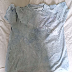 Ljusblå STWD t-shirt från Pull&Bear - Ljusblå t-shirt från Pull&Bear STWD med trycket 'Surviving these weird days' på bröstet. T-shirten har en avslappnad passform, rund hals och är tillverkad i mjuk bomull. Perfekt för dig som gillar chill och stilrena plagg med coolt statement.